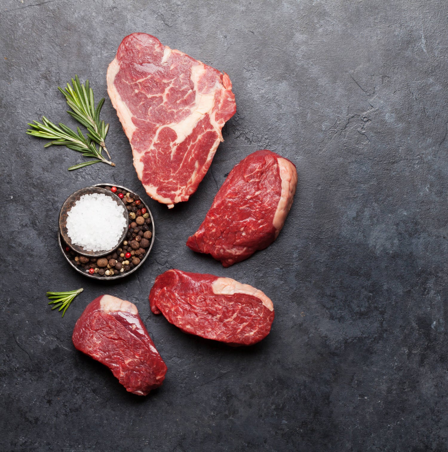 Raw steak cuts on a blackboard with fresh herbs and spices